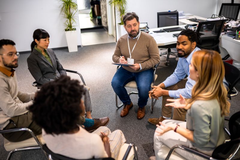 A diverse group of six adults sits in a circle in an office, engaged in conversation. 