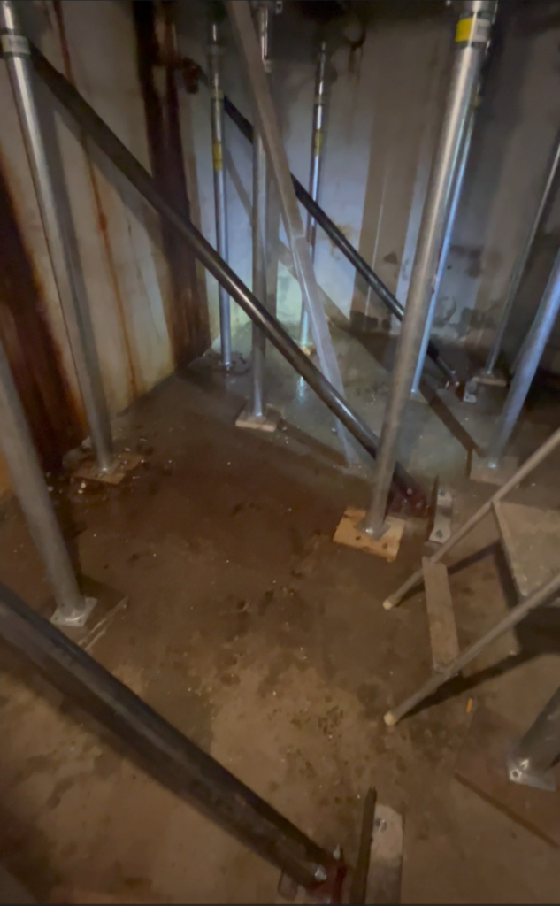 Close-up of the interior corner of an underground sidewalk vault with peeling concrete, staining, and obsolete utility wiring before removal and repair.